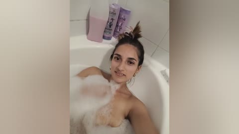 Media: Video of a young, topless woman with medium brown skin and dark hair in a bun, partially submerged in a white bathtub filled with bubbles. Behind her, a lavender shampoo bottle and pink conditioner sit on the tub's edge.