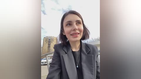 Media: Video of a smiling young woman with shoulder-length dark hair, wearing a gray blazer over a black top, standing outdoors in an urban setting with modern buildings and a cloudy sky.