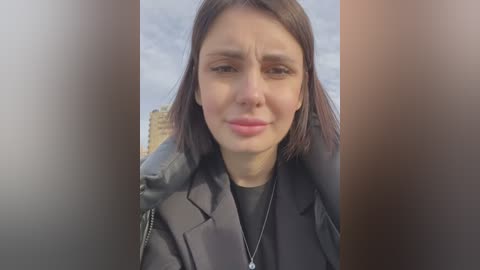 Media: Video of a young woman with light skin, straight brown hair, and a serious expression, wearing a black coat, standing outdoors with a blurred urban skyline in the background.