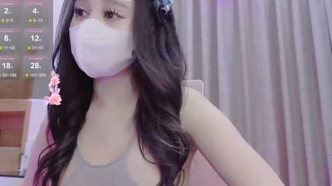 Media: Video of a young Asian woman with long, wavy black hair and a pink flower accessory, wearing a white surgical mask, gray tank top, and standing indoors against a light-colored curtain background.