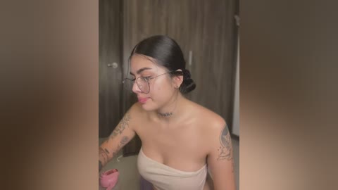 Media: Video of a young woman with light skin, long black hair in a bun, wearing glasses, and a strapless top. She has multiple tattoos on her arms, standing in a dimly lit room with wooden doors.