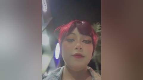 Media: A video of an Asian woman with short, vibrant red hair and heavy makeup, including red lipstick and dark eyeliner, wearing a light jacket. The background is blurry, suggesting an urban setting with streetlights.