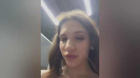 Media: A blurry video of a young woman with light skin and long, wavy brown hair, wearing a pink top and minimal makeup, standing in an indoor setting with fluorescent lights.