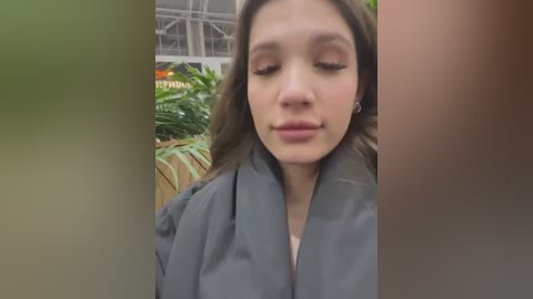 Media: A video of a young woman with long brown hair and closed eyes, wearing a gray coat, standing in a greenhouse with lush green plants and a blurred background.