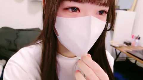 Media: Video of an Asian woman with long, straight brown hair and bangs, wearing a white face mask and white shirt, holding mask strings, in a cluttered indoor setting with a green couch, table, and various items in the background.