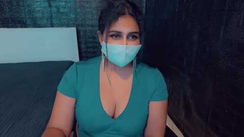 Media: Video of a curvy woman with dark hair, wearing a teal V-neck top, blue face mask, and dark makeup, sitting on a bed against a dark wall, with a neutral background.