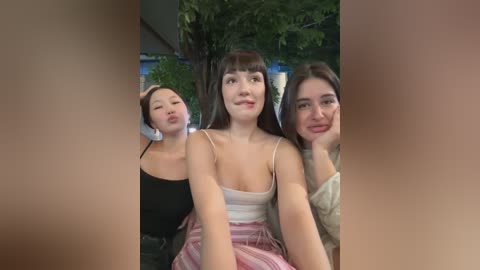 Media: Video of three young women with straight, dark hair, smiling and leaning against each other, wearing casual clothing. Background includes green foliage and a building.