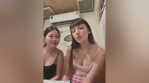 Media: Video of two young Asian women with straight black hair, wearing casual tank tops, standing in a small, dimly lit room with posters on the wall.