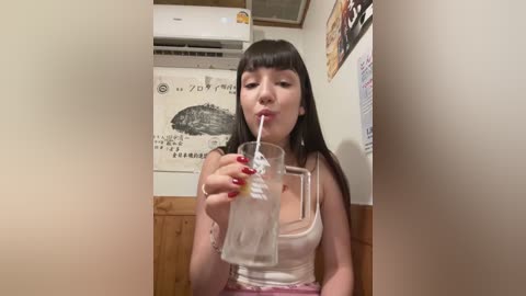 Media: Video of an Asian woman with straight black hair and light skin, wearing a white tank top and pink shorts, drinking from a clear glass, in a cozy, cluttered room with wooden walls and a mounted air conditioner.