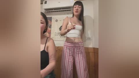 Media: Video of an Asian woman in a white crop top and pink striped pajama pants, with another woman in black tank top and green pants partially visible, in a cozy indoor setting with wooden paneling and an air conditioner.