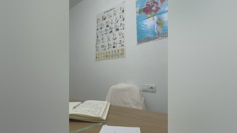 Media: Video of a small, sparsely furnished classroom with white walls, a map of Europe, and a chalkboard displaying a phonics chart. A desk holds an open notebook, and a white chair is partially visible.