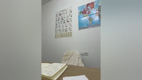 Media: A video of a small, simple classroom with a beige wall, a child's alphabet poster, a world map, a wooden desk, and an open notebook with handwritten notes.