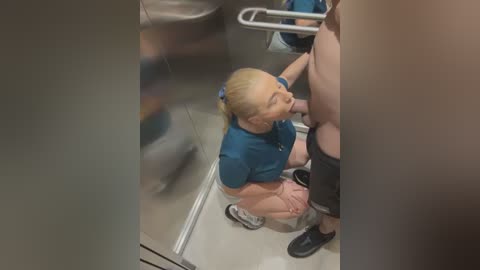 Media: Video of a blonde-haired woman in a blue uniform and pink pants squatting to assist a male with a black leg brace in a public restroom.