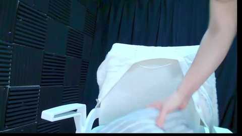 Media: A video of a person in a white hospital gown, adjusting a plastic bag on a hospital bed. The background features dark, textured wall panels and a curtain.