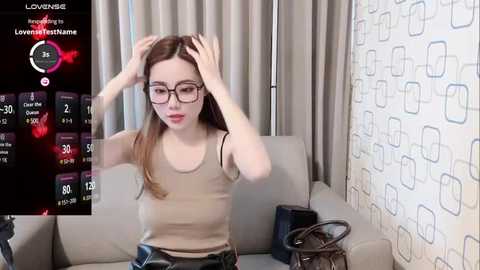 Media: Video of a young woman with glasses, fair skin, and long brown hair, adjusting her hair on a beige couch in a modern living room with patterned wallpaper.