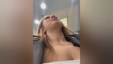 Media: Video of a blonde woman with fair skin, wearing a black jacket, leaning back with eyes closed, taken from a low angle. Background includes modern, minimalist interior with beige walls and recessed lighting.