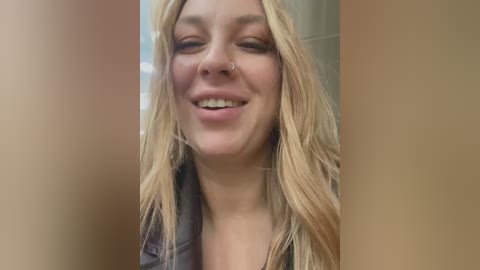 Media: Video of a smiling, fair-skinned, blonde woman with long, wavy hair, wearing a gray jacket, set against a blurred, beige background.