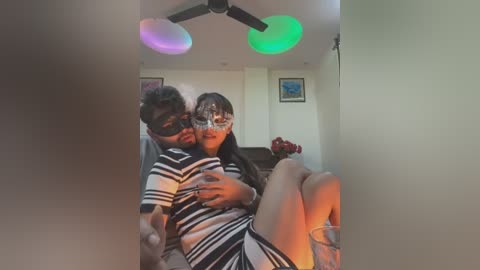 Media: Video of a couple in a dimly lit room; woman with dark hair, wearing a black and white striped dress and silver mask, sitting on a man's lap, who's dressed in a striped shirt.