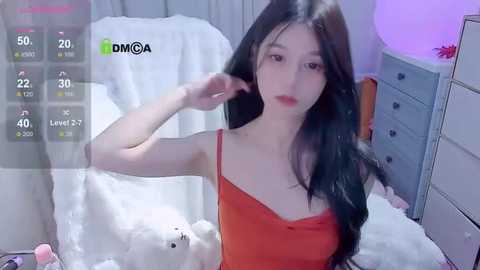 Media: Video of an East Asian woman with long black hair, fair skin, wearing a red camisole, in a cozy, softly lit bedroom with white bedding, a blue dresser, and a fluffy white rug.