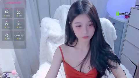 Media: Video of an East Asian woman with long, straight black hair, wearing a red camisole, sitting on a white furry chair in a minimalist, softly lit room.