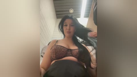 Media: Video of a curvy Latina woman with long black hair and medium skin tone, wearing a leopard-print bra, lying on a bed, in a dimly lit, narrow hallway with white tiled walls and a black ceiling.