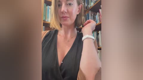 Media: A video of a woman with short blonde hair, wearing a black sleeveless top and a white bracelet, standing in front of bookshelves filled with books.