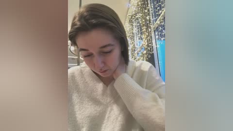 Media: Video of a young woman with light skin and straight, dark brown hair, wearing a white knit sweater, looking down, touching her neck, in a dimly lit room with green curtains and blue light.