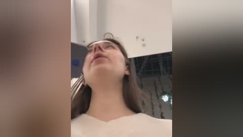 Media: Video of a woman with long, straight brown hair, wearing a white shirt, looking upwards, possibly in an auditorium with dim lighting and visible seating.