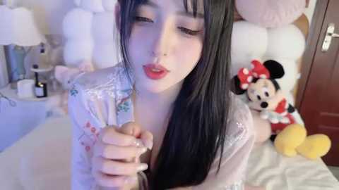 Media: A video of a fair-skinned, slender Asian woman with long black hair, wearing a white floral robe, kissing her hand. The background features a bed with plush toys, including Mickey Mouse. The room has a soft, pastel color scheme.