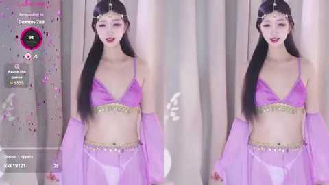 Media: Video of a pale-skinned East Asian woman with long black hair, wearing a sheer purple belly dance outfit, with a gold belt and matching headpiece, posing in front of a glittery backdrop.