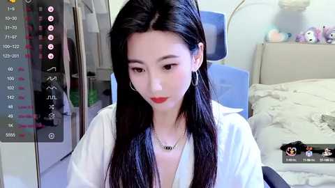 Media: A video of an East Asian woman with long, straight black hair, fair skin, and red lipstick, wearing a white robe and silver necklace, sitting on a bed in a bedroom.