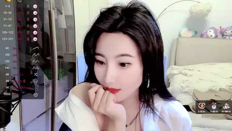 Media: A video of a young Asian woman with long black hair and fair skin, wearing a white shirt, playing with her mouth in a bedroom. The room is light and has a bed and stuffed toys.