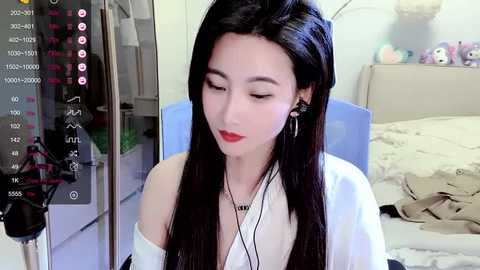 Media: A video of a young Asian woman with long black hair, fair skin, and red lipstick, wearing a white off-shoulder blouse, sitting in a bedroom with a bed, toys, and a mirror.