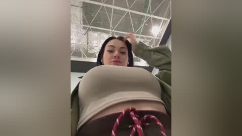 Media: Video of a young woman with dark hair, wearing a beige crop top and green jacket, standing in an industrial space with a white lattice ceiling and exposed pipes.