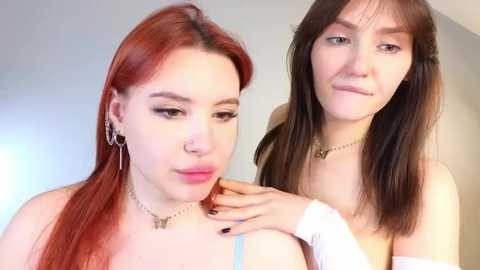 Media: Video of two young women with fair skin, one with red hair, the other with brown hair, both wearing minimal makeup and gold jewelry, touching each other's shoulders.