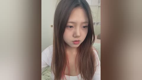 Media: A video of a young East Asian girl with long brown hair, light skin, and a neutral expression, wearing a white top, in a dimly lit room with a beige wall and a window with blinds.