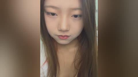 Media: Video of a young Asian woman with long, straight brown hair and light skin, wearing minimal makeup, standing indoors with blurred background.