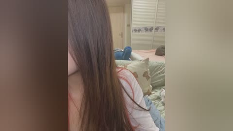 Media: Video of a woman's long brown hair partially obscuring her face, standing in a messy bedroom with a white bed, blue pillow, and closed blinds.