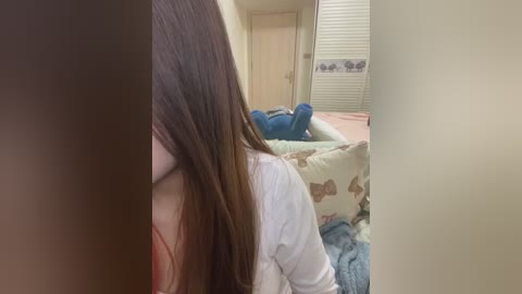 Media: Video of a young woman with long, straight brown hair, wearing a white shirt, lying on a bed with a patterned duvet in a modestly decorated bedroom.