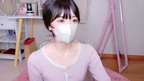 Media: Video of an East Asian woman with short dark hair, wearing a white face mask and light pink cardigan, sitting on a pink rug in a minimalistic room with white walls, wooden floor, and pink mirror.