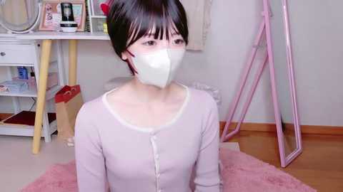 Media: Video of a young East Asian woman with short black hair, wearing a light pink cardigan, white mask, and sitting on a pink rug. Background includes a white desk, pink chair, and a mirror.