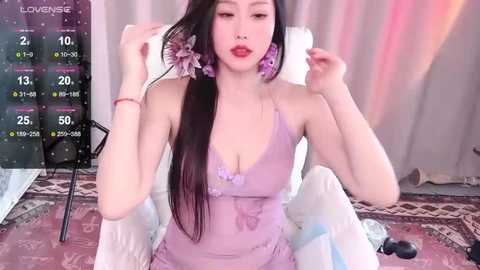 Media: A video of an Asian woman with long black hair, wearing a purple lace dress, sitting on a chair. She has large purple earrings and is adjusting her hair. Background includes a bed with white sheets, a digital screen displaying a video game, and a patterned rug.