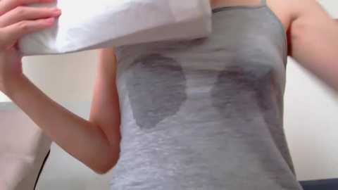 Media: Video of a fair-skinned woman with medium build, wearing a grey tank top with a large, abstract black design, holding a white towel over her chest.