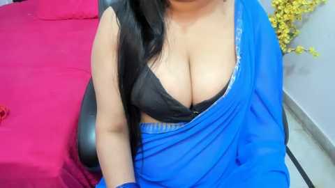 Media: A video of a South Asian woman with long black hair, wearing a blue sari and a black bra, revealing cleavage, sitting on a pink bed with yellow flowers in the background.