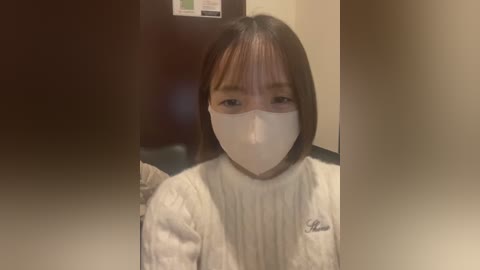 Media: A video of an East Asian woman with short, straight black hair and bangs, wearing a white surgical mask and a white lab coat, seated indoors against a blurred background.