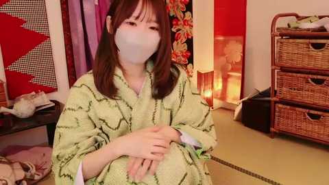 Media: A video of a woman with shoulder-length brown hair, wearing a light green kimono with a floral pattern, seated on the floor. The background features traditional Japanese decor, including a red wall, a woven basket, and a colorful screen.