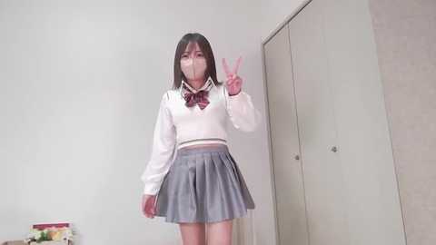 Media: Video of an Asian woman with long black hair in a schoolgirl outfit: white sweater, plaid bow tie, pleated gray skirt. She makes a peace sign in a minimalistic, beige-walled room.