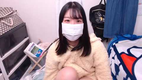 Media: Video of an Asian woman with straight black hair, wearing a white mask, beige fuzzy sweater, and sitting on a bed. Background includes a blue backpack, white patterned blanket, and a grey and white patterned pillow.