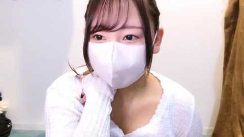 Media: Video of an East Asian woman with straight, dark hair, wearing a white face mask and white sweater, flossing her teeth in a bathroom with light walls and a brown curtain.