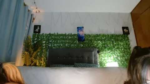 Media: Video of a cozy, dimly lit bedroom with a grey tufted headboard, green faux-plant wall, yellow potted plant, blue curtains, and two framed portraits.